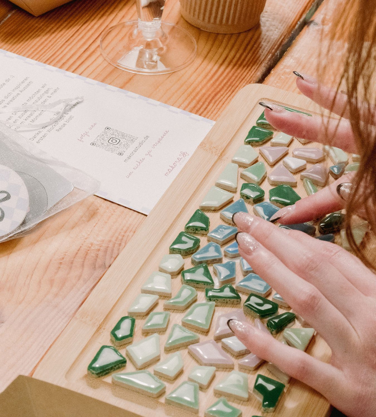 Hände legen grüne, blaue und rosa Mosaiksteine auf einem Holzbrett zurecht